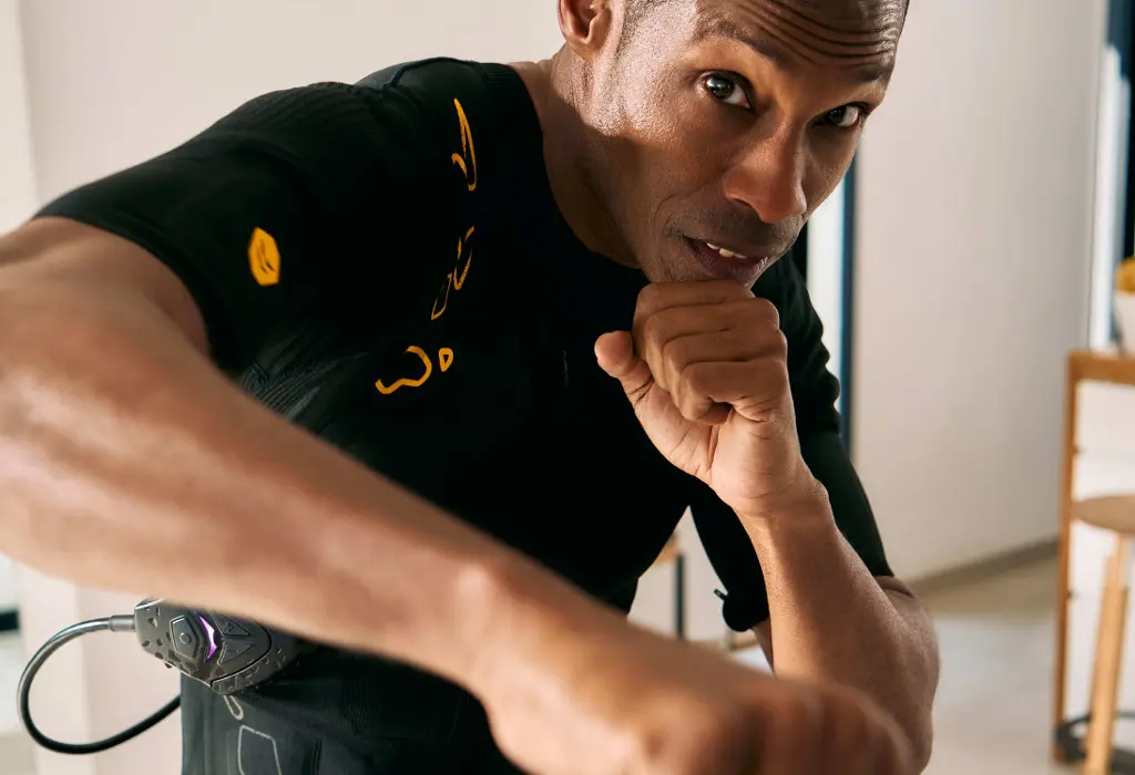A person in a SYMBIONT EMS suit performs boxing movements with a focused facial expression. The image represents an advanced training experience.