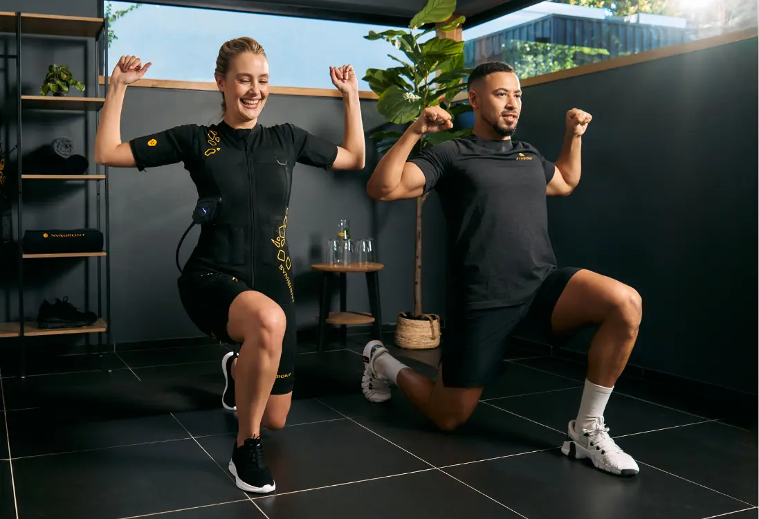 A woman in an EMS suit is performing exercises under the guidance of a professional trainer to ensure effective and personalized training. She is working out in a minimally furnished, light-filled room.